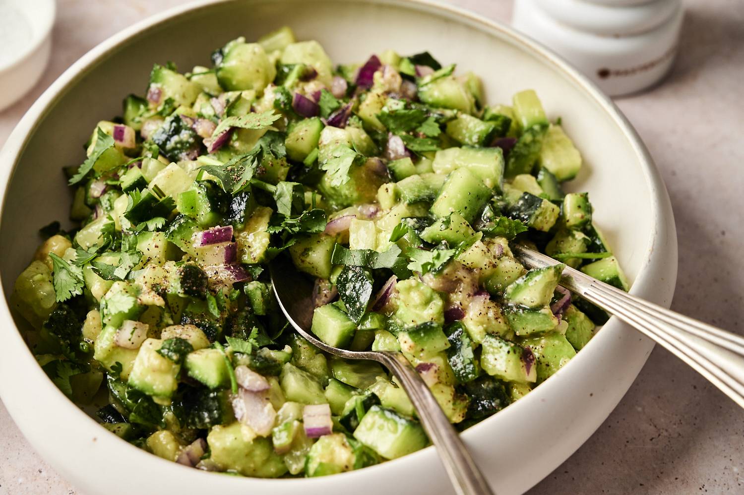 Close-up of avocado cucumber salsa in a white bowl, mixed with red onion, fresh cilantro, and seasonings, served with a spoon for easy scooping.