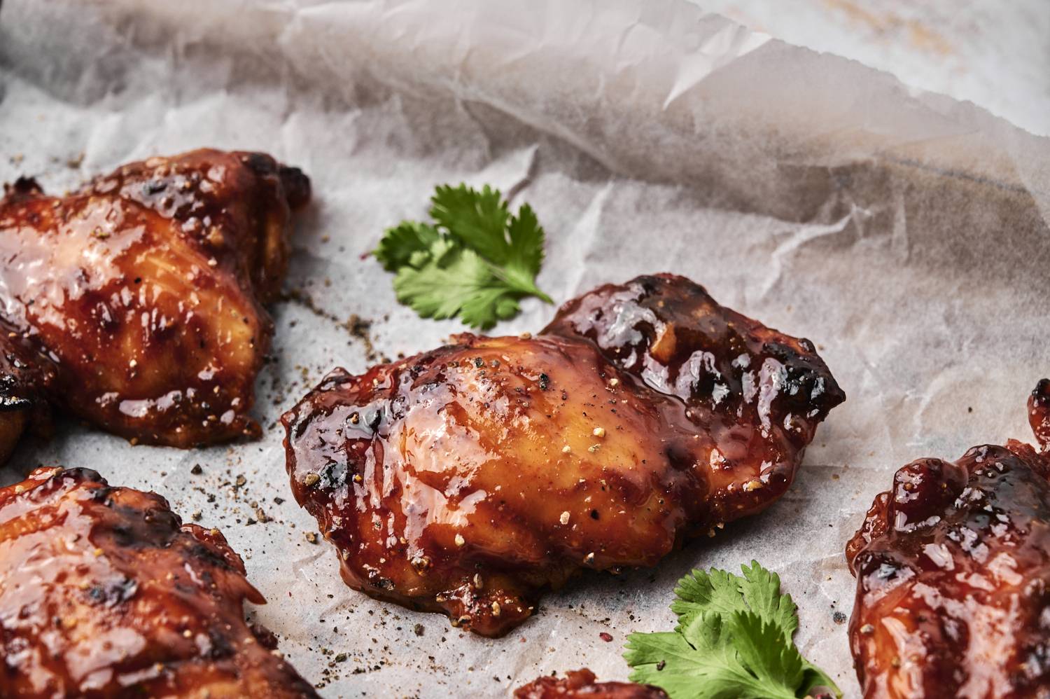 Close-up of juicy baked BBQ chicken thighs on a parchment-lined baking sheet, coated in a glossy, caramelized barbecue glaze and garnished with fresh cilantro.