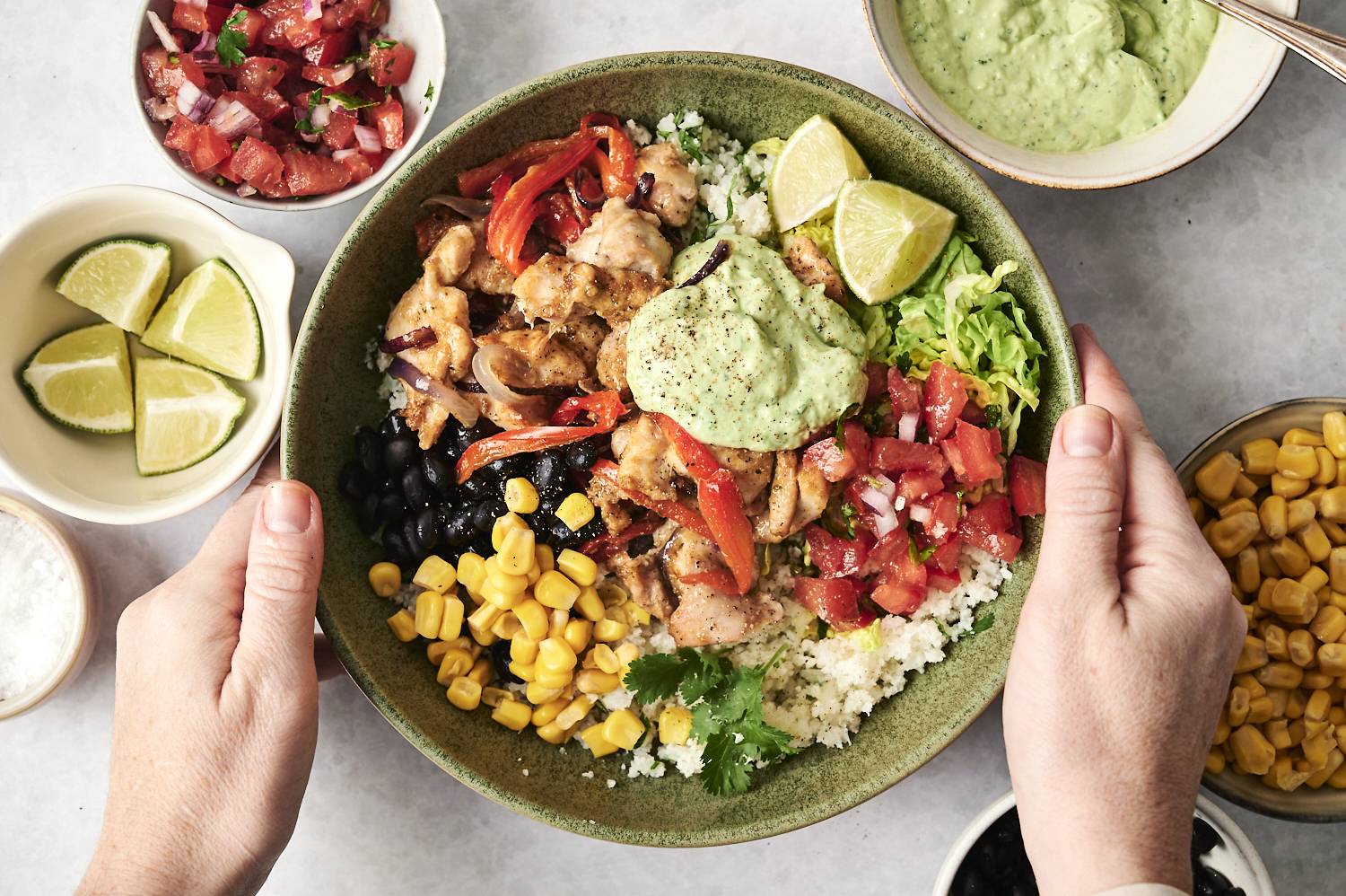 Hands holding a bowl filled with roasted chicken, cauliflower rice, black beans, corn, pico de gallo, lettuce, and creamy green sauce, surrounded by fresh ingredients.