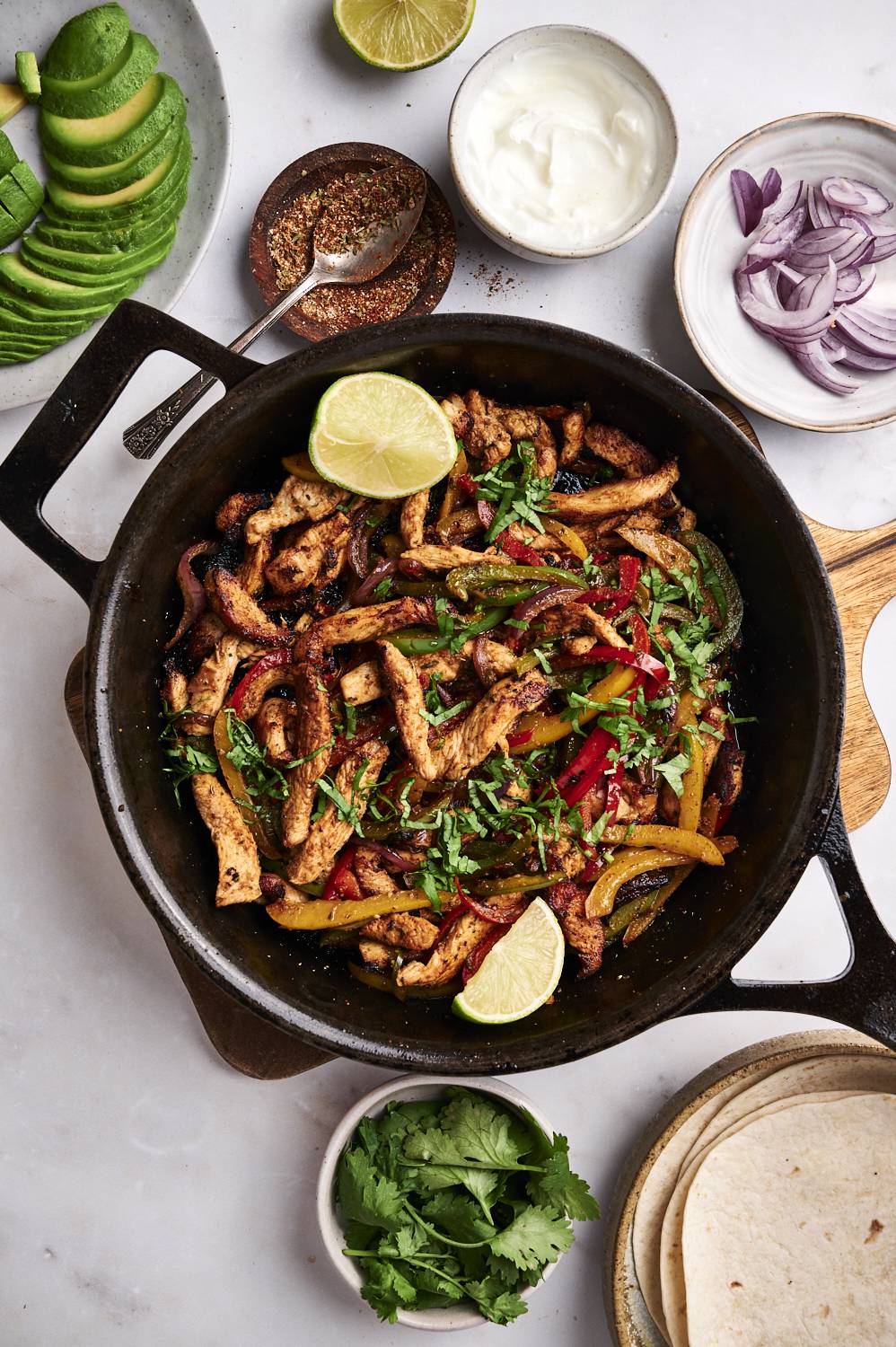 Skillet chicken fajitas served with cilantro, sour cream, lime wedges, flour tortillas, and sliced avocado.