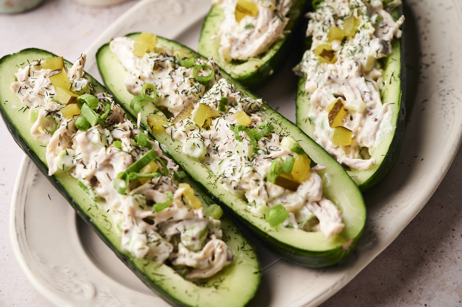 Halved cucumbers hollowed out and filled with creamy chicken salad, topped with chopped pickles, sliced green onions, and a sprinkle of dried dill, served on a white oval plate.