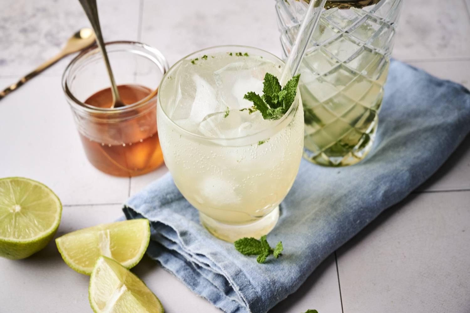 A refreshing cocktail with ice and mint leaves, accompanied by lime halves and a jar of honey on a denim cloth.