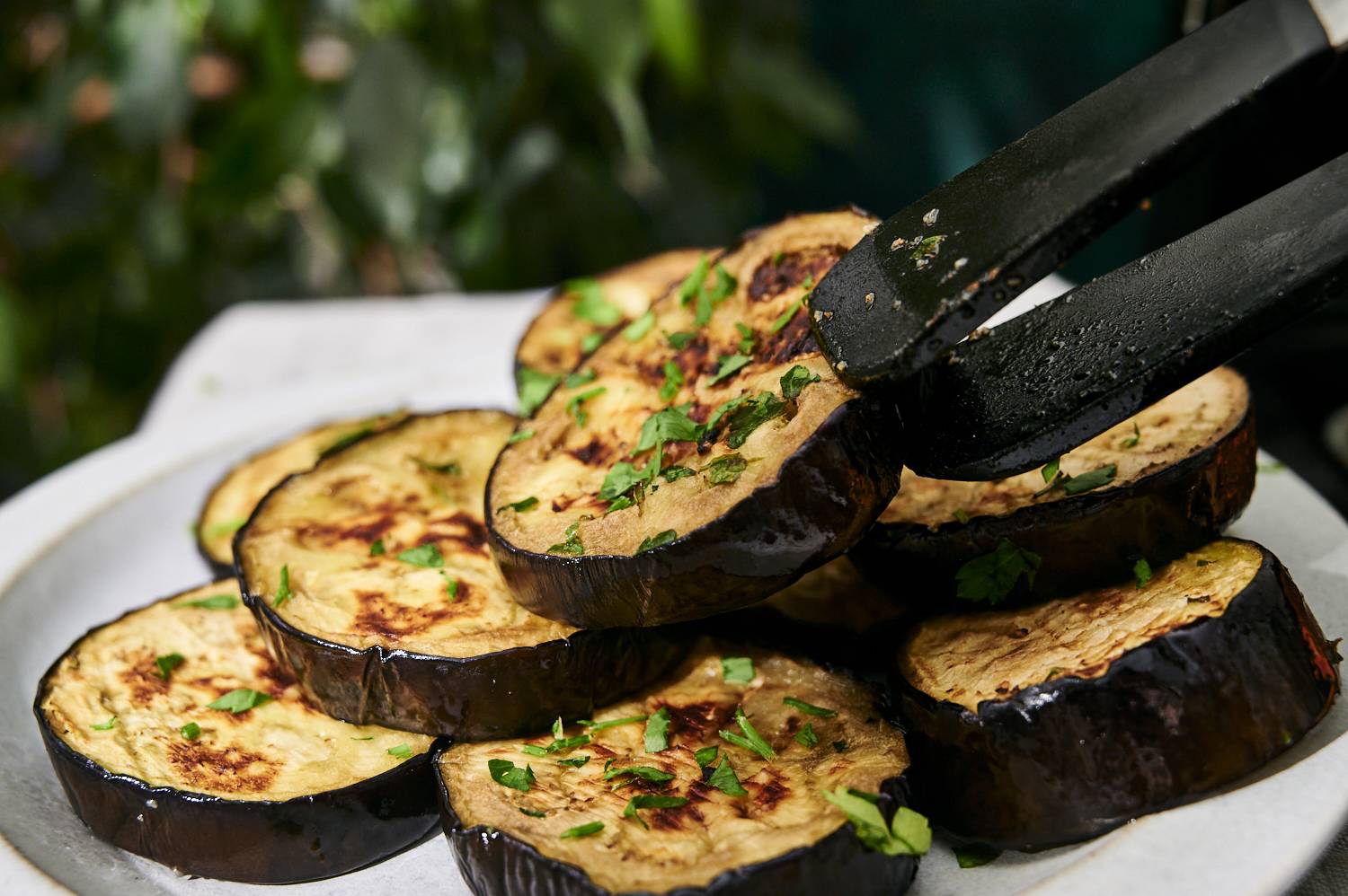 A plate of grilled eggplant slices garnished with fresh herbs, being lifted with tongs, set against a blurred green background.