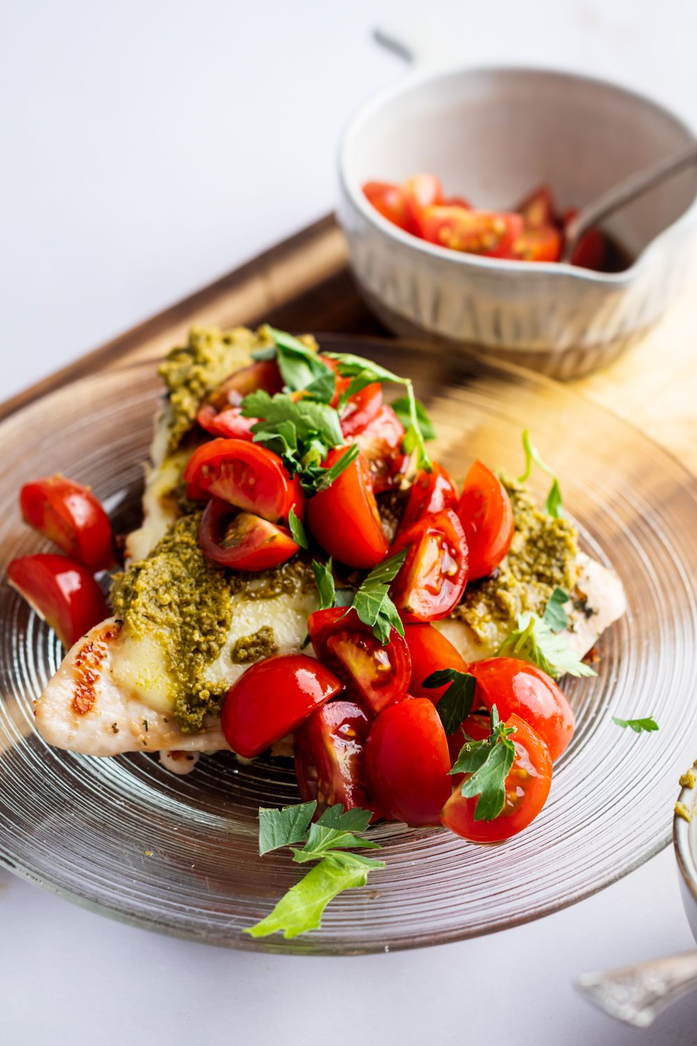 Grilled chicken breast served with tomato salad, melted mozzarella cheese, and pesto on a glass plate with tomatoes on the side.