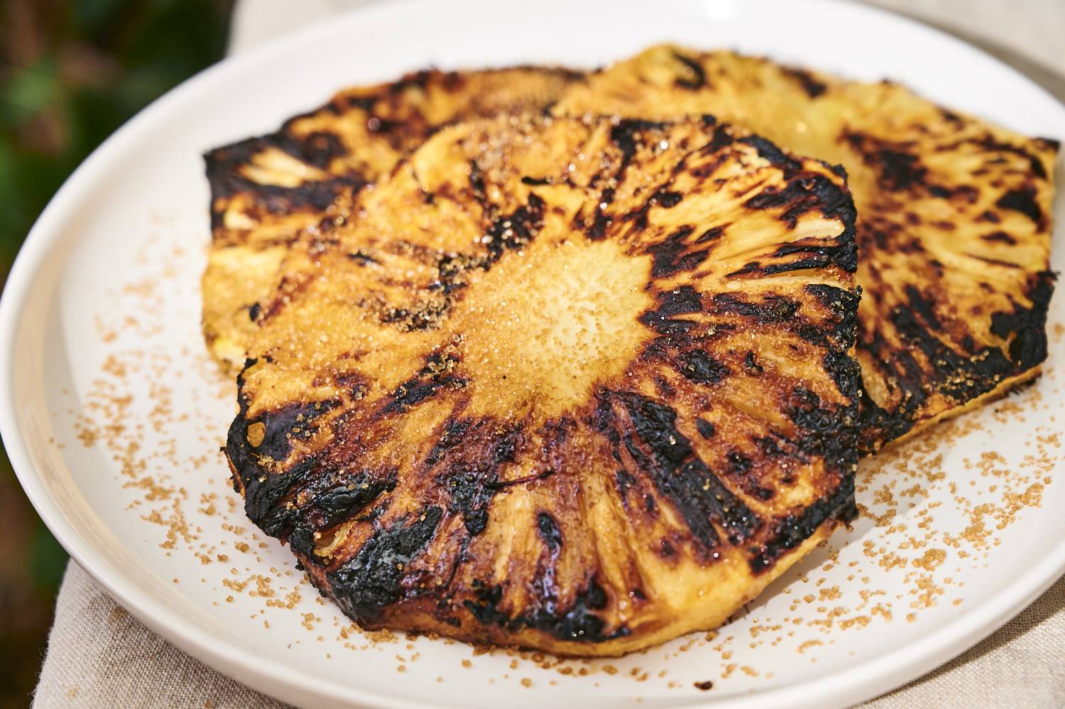 Grilled pineapple slices with caramelized edges, sprinkled with brown sugar, on a white plate. Warm and inviting presentation.