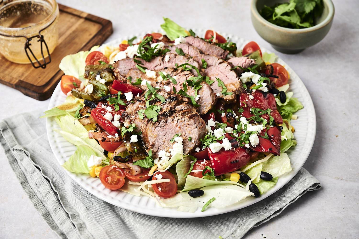 A vibrant salad topped with grilled beef, cherry tomatoes, peppers, cilantro, and feta, served on a plate with a jar of dressing.
