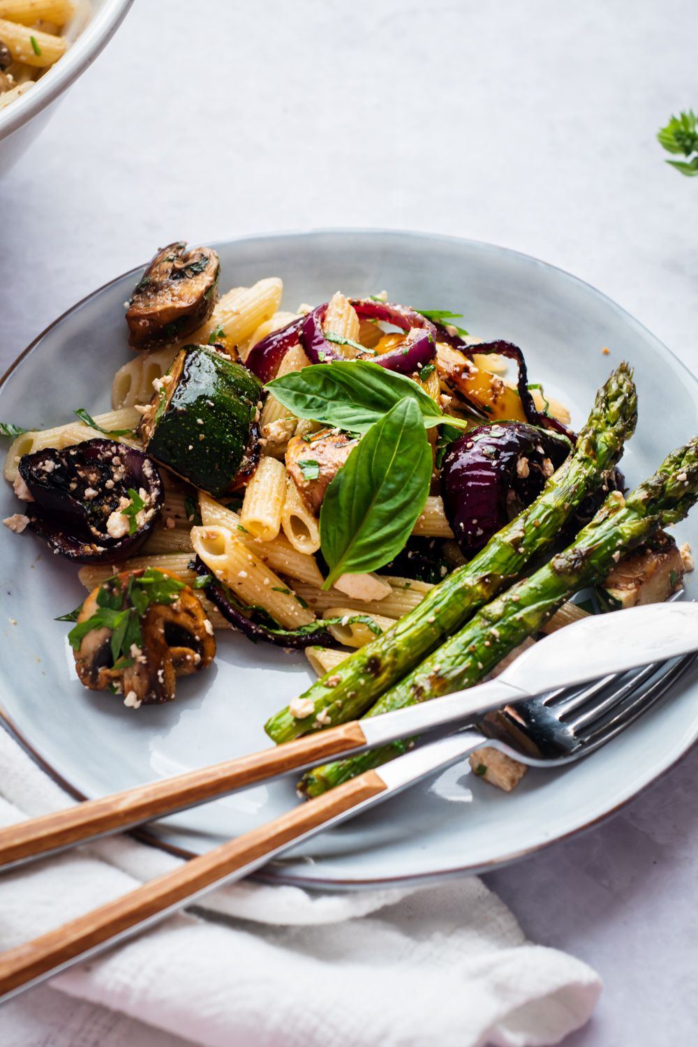Pasta salad with grilled vegetables, feta cheese, basil, and parsley on a plate with a fork and knife.