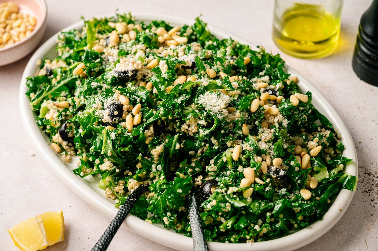 A vibrant kale salad on a white platter topped with quinoa, pine nuts, and grated cheese. Olive oil and lemon wedge sit nearby, evoking freshness.