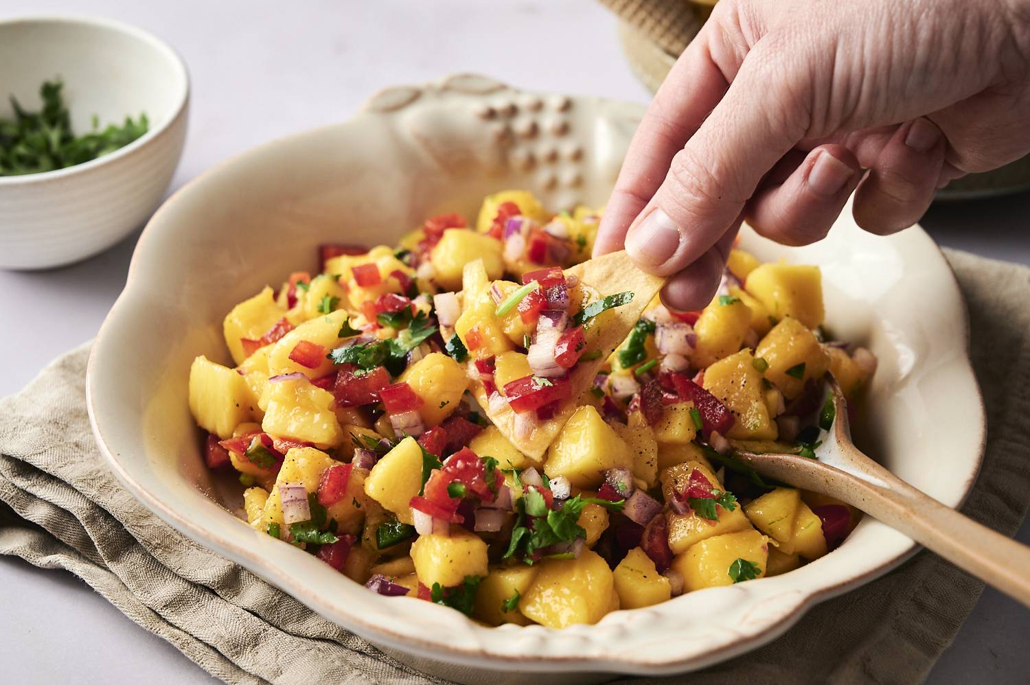 Hand dipping a tortilla chip into a bowl of fresh mango salsa made with diced mango, red bell pepper, red onion, jalapeño, and cilantro.