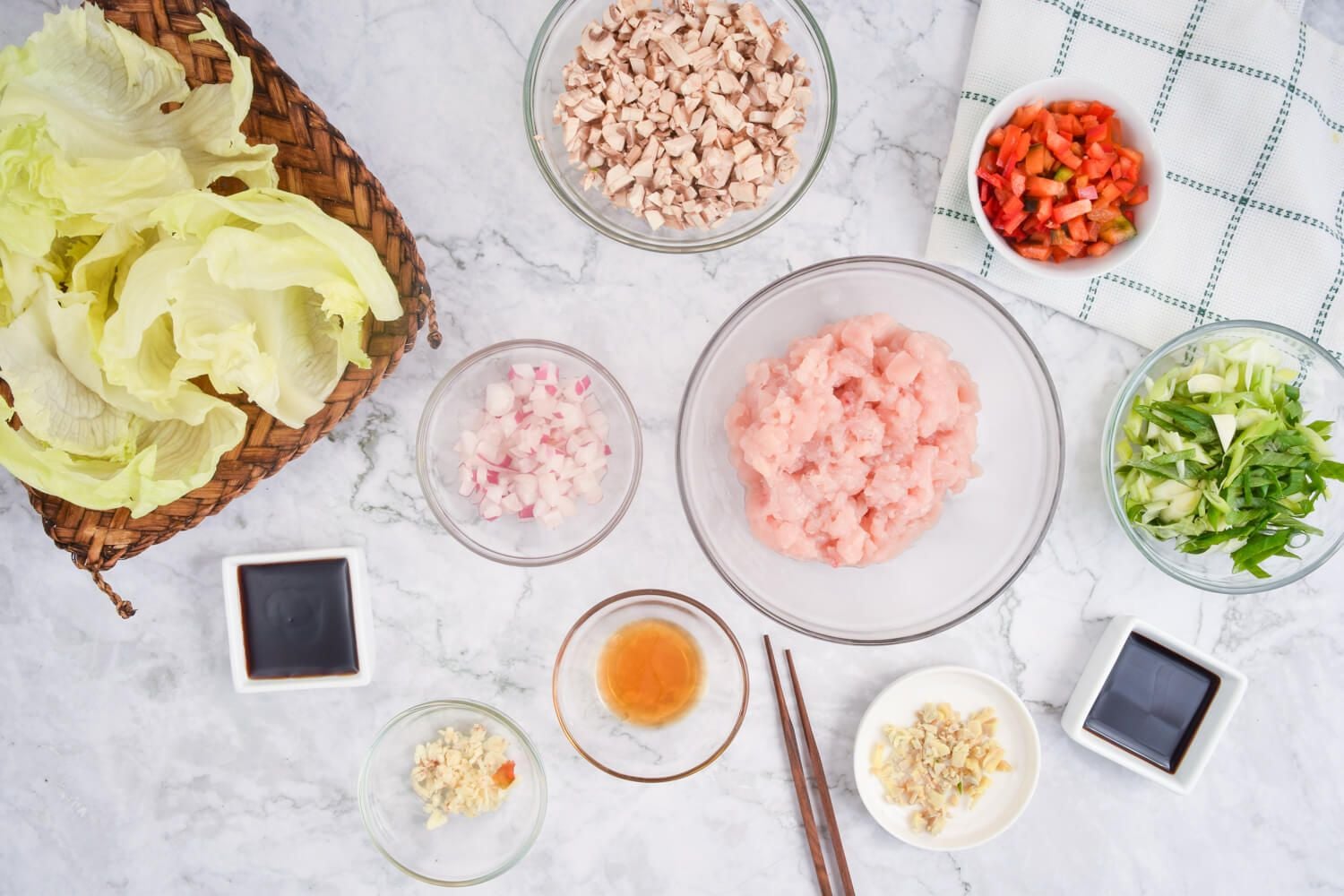 Ingredients for chicken lettuce wraps on a marble background including chicken, lettuce, mushrooms, and red peppers.