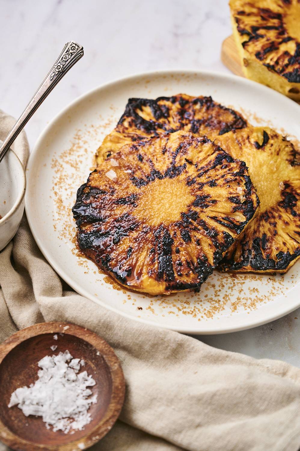 Grilled pineapple slices on a white plate with a sprinkle of sugar, alongside a bowl of flaky sea salt and a silver spoon.