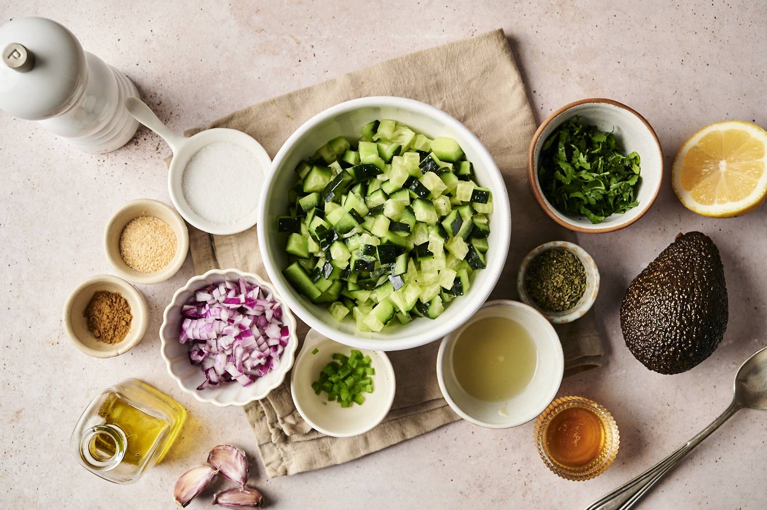 Overhead view of diced cucumbers in a white bowl surrounded by ingredients for avocado cucumber salsa, including red onion, jalapeño, cilantro, garlic, lemon, avocado, spices, olive oil, and salt on a light surface.