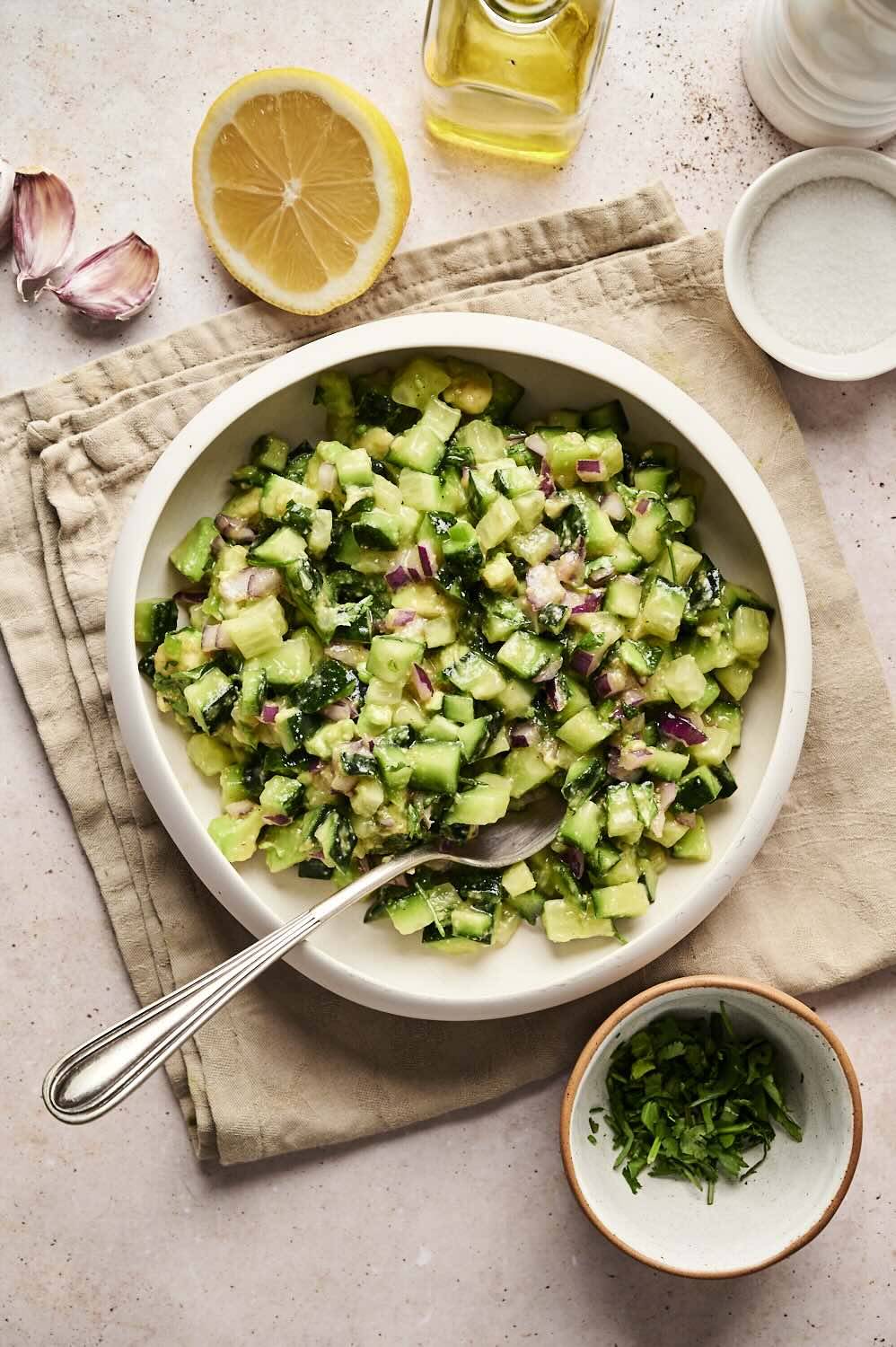 Overhead view of avocado cucumber salsa in a white bowl with a spoon, surrounded by lemon, garlic, olive oil, salt, and fresh cilantro on a light surface.