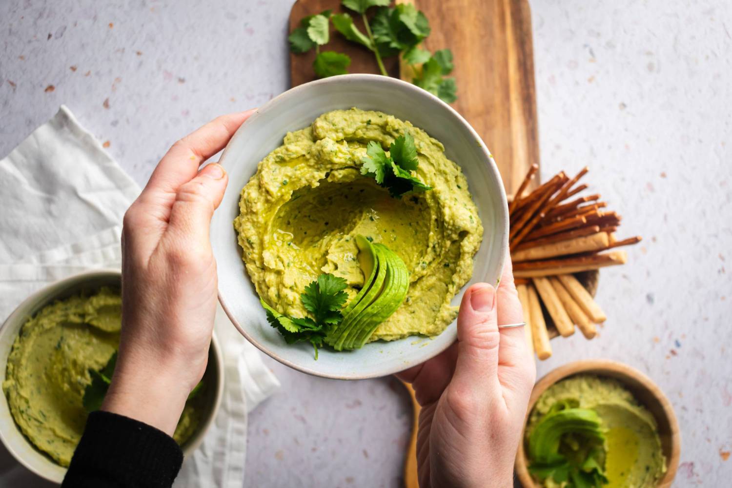 Creamy avocado hummus with chickepas, tahini, avocado, lemon juice, and cilantro in a bowl being held by two hands.