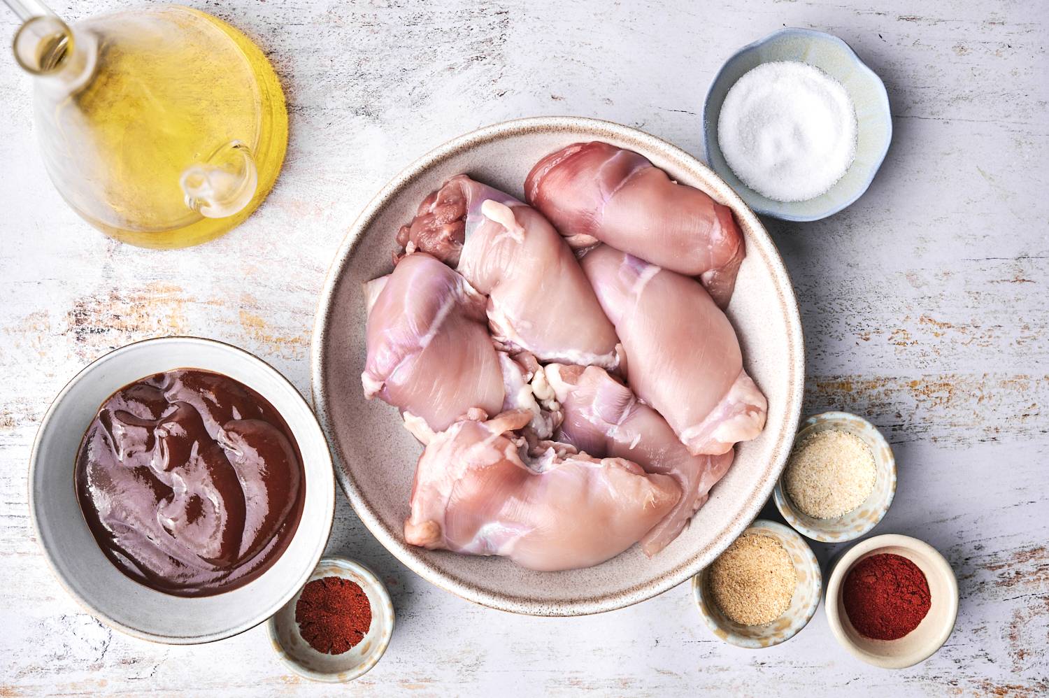 A bowl of raw chicken thighs surrounded by spices, olive oil, and a dark sauce on a rustic wooden surface.