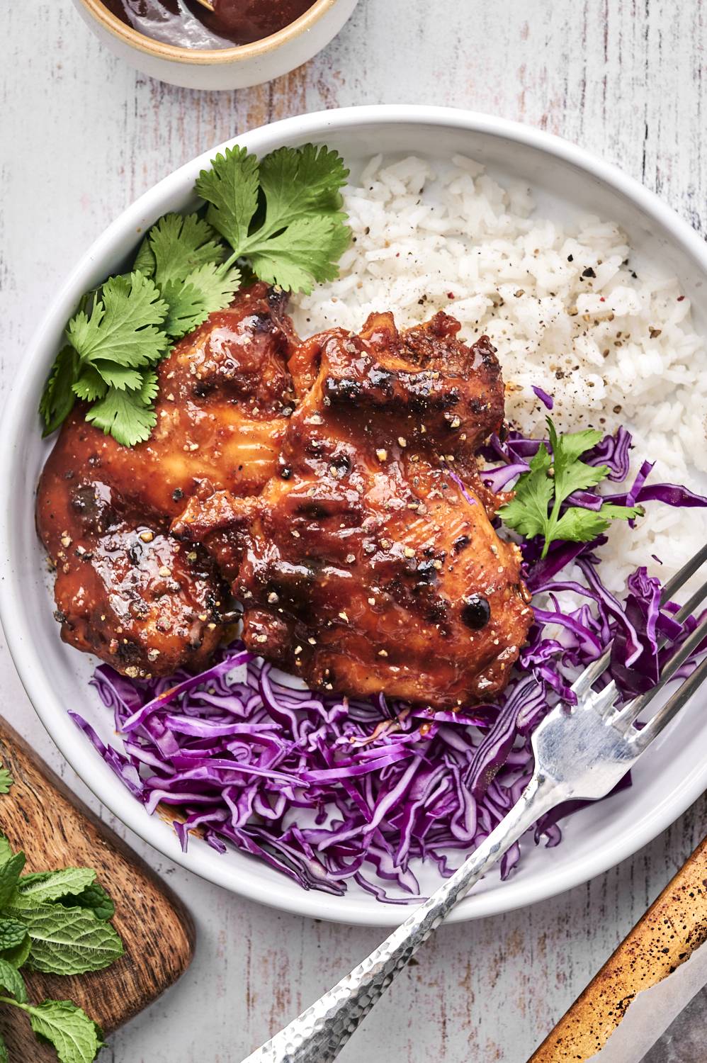 A bowl of sticky, glazed chicken over fluffy white rice, topped with purple cabbage and fresh cilantro, against a rustic backdrop.