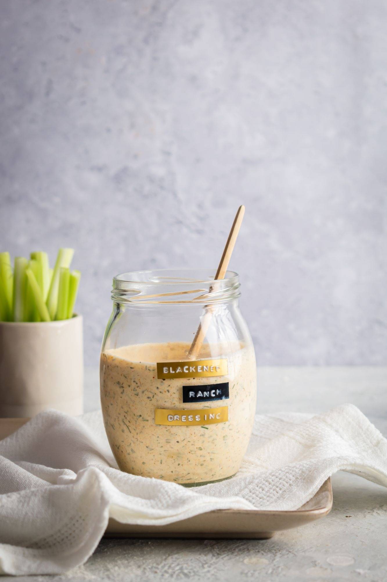 A glass jar labeled “Blackened Ranch Dressing” filled with creamy dressing speckled with herbs, with a wooden spoon inside, set on a white cloth with celery sticks in the background.