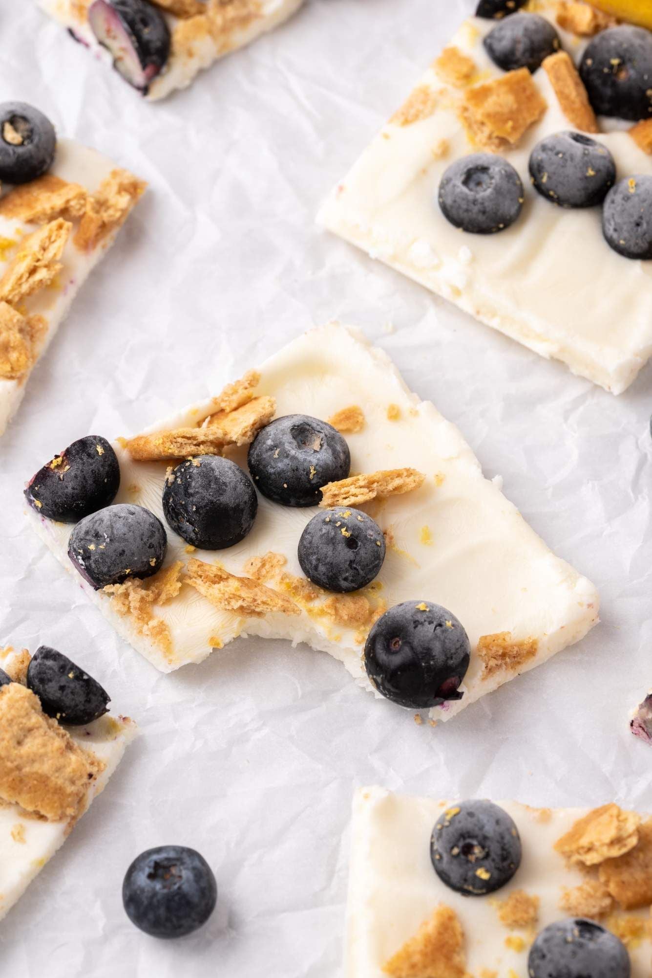 Frozen yogurt bark pieces with fresh blueberries, lemon zest, and graham crackers on parchment paper.