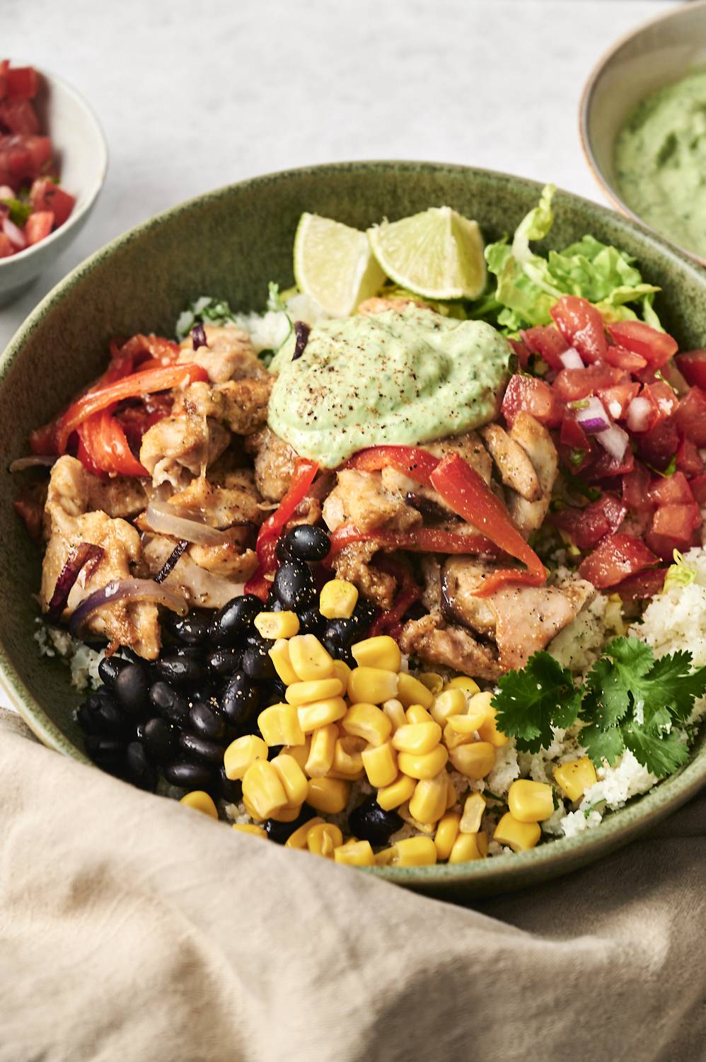 A vibrant chicken and cauliflower rice bowl topped with black beans, corn, pico de gallo, avocado sauce, and lime wedges.