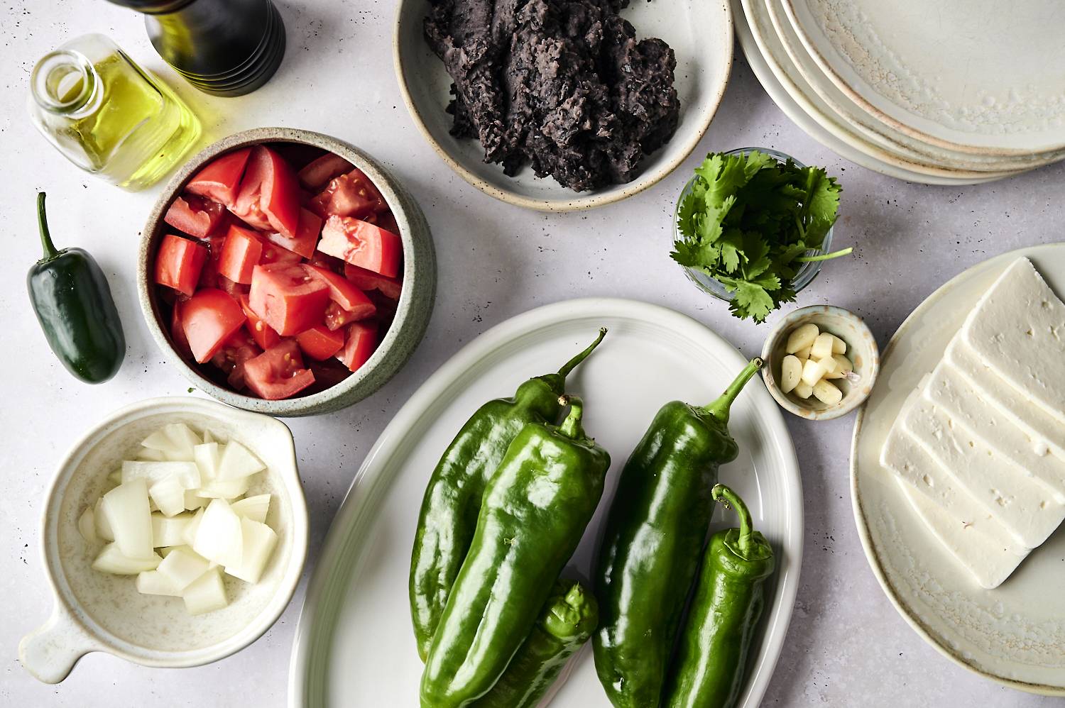 Various ingredients for preparing chiles rellenos are laid out on a countertop. There are green poblano peppers, diced tomatoes, chopped onions, a bunch of fresh cilantro, garlic, black beans, and slices of cheese. A bottle of olive oil sits to the side with a bowl of jalapeño.
