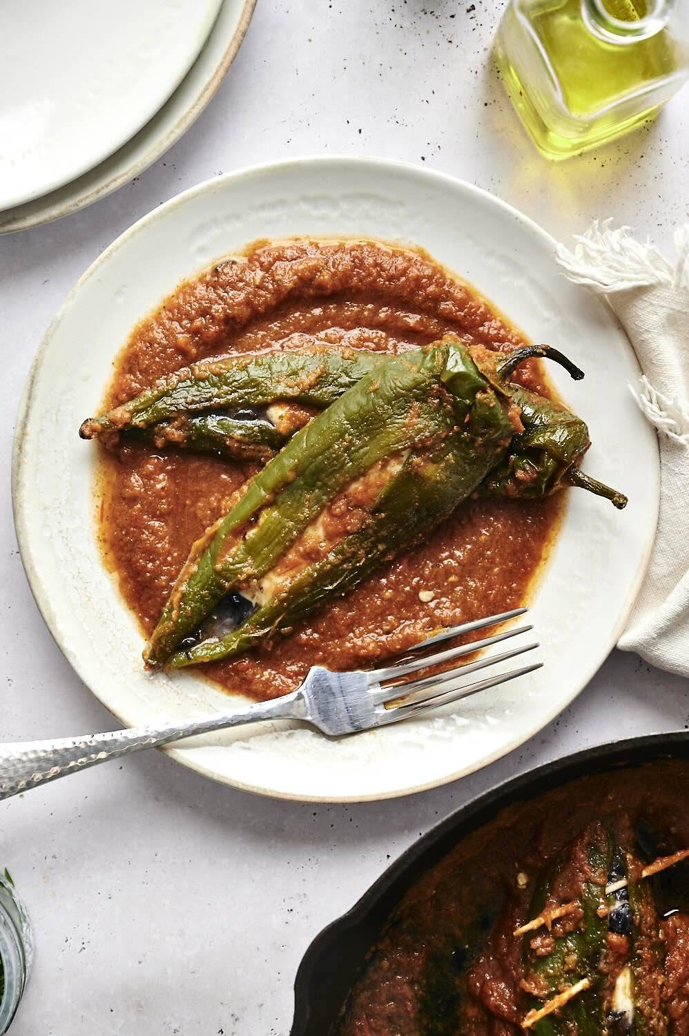 A plate of roasted green chile peppers served on a bed of red sauce, garnished with a fork and accompanied by a bottle of olive oil in the background.