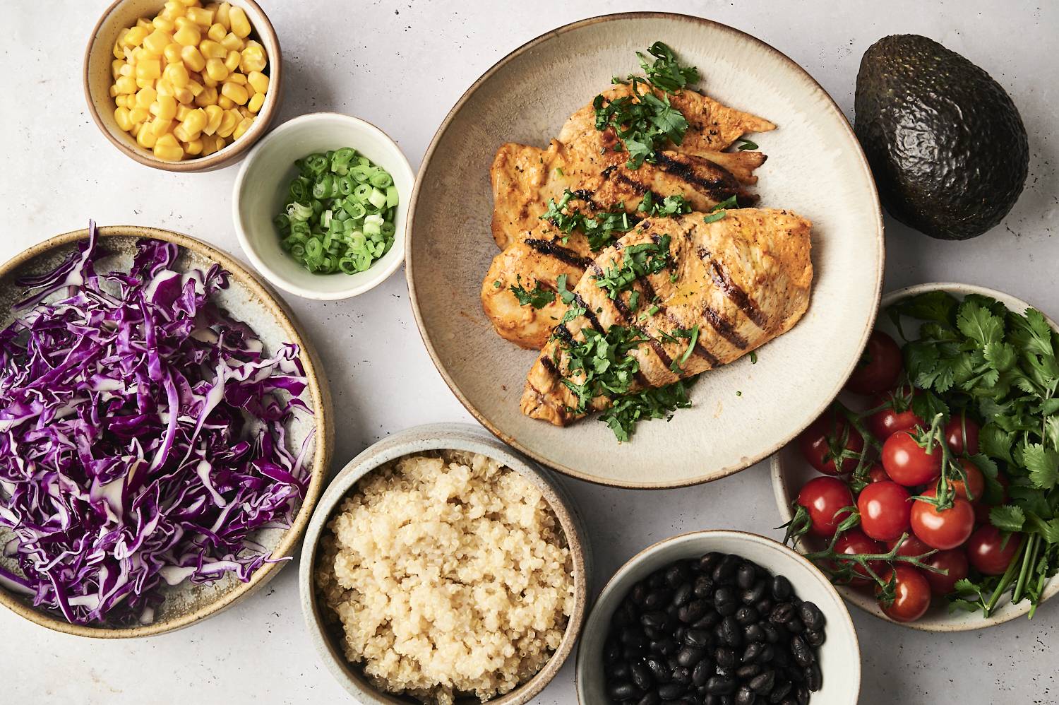 Grilled chicken breast garnished with cilantro, served with quinoa, black beans, cherry tomatoes, corn, shredded red cabbage, and sliced green onions, alongside an avocado and cilantro.