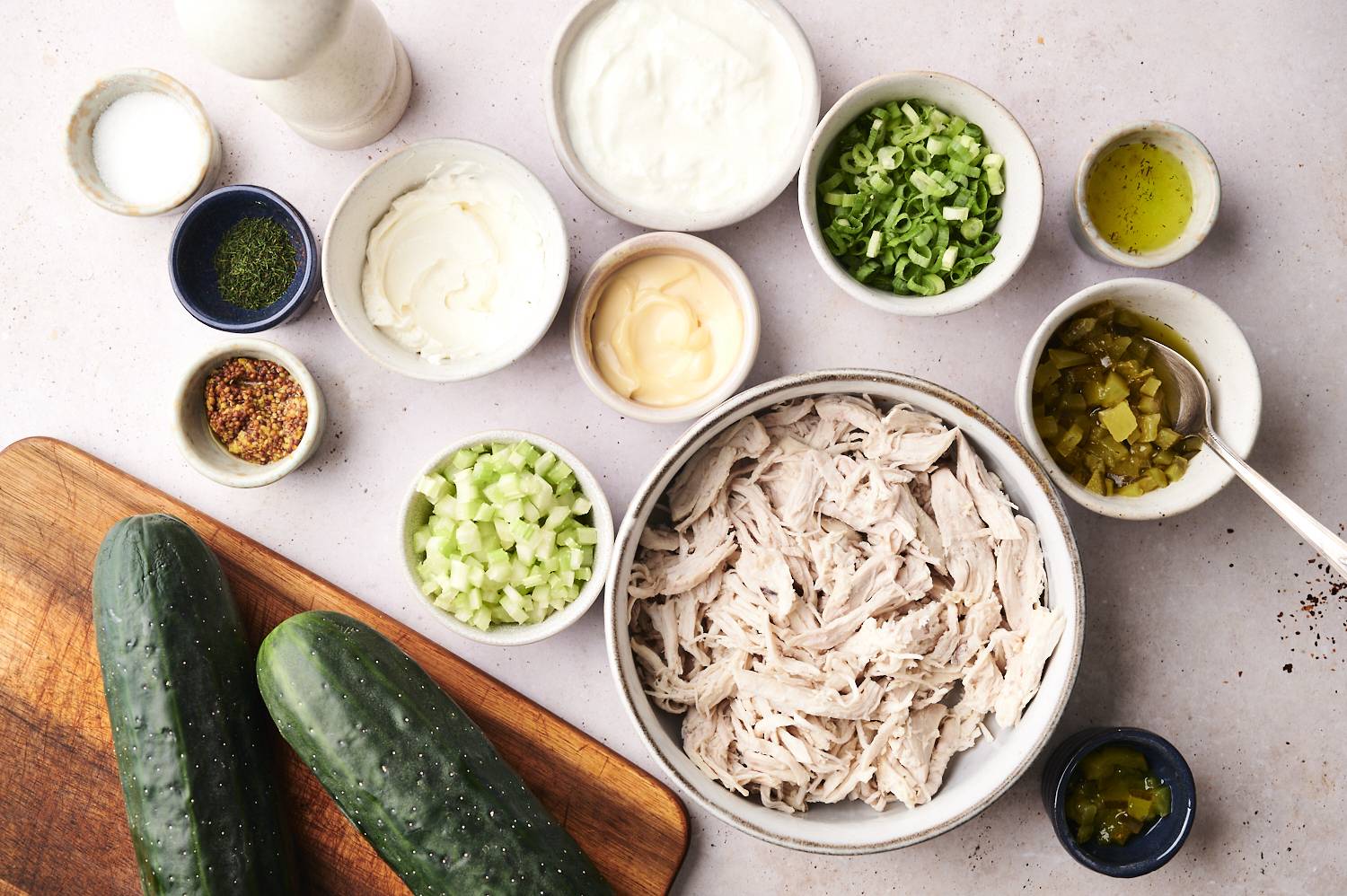 Overhead view of ingredients for dill pickle cucumber boats, including shredded chicken, diced celery, chopped green onions, diced pickles, cucumbers, mayonnaise, Greek yogurt, cream cheese, whole grain mustard, dried dill, pickle juice, salt, and pepper, all arranged in bowls on a light surface with a wooden cutting board. 