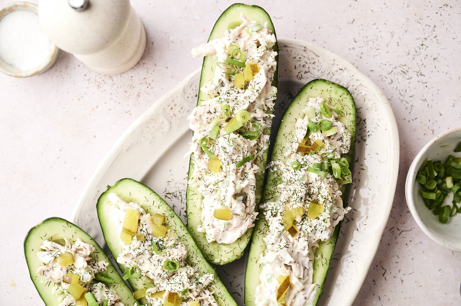 Close-up view of dill pickle cucumber boats on a white serving platter, each cucumber half filled with creamy shredded chicken salad, topped with chopped pickles, sliced green onions, and sprinkled with dried dill. A small bowl of extra green onions sits nearby.