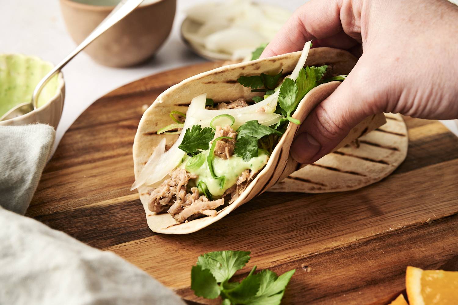 A hand holding a soft taco filled with shredded pork carnitas, fresh cilantro, sliced onions, and creamy green sauce on a grilled tortilla, set on a wooden board.