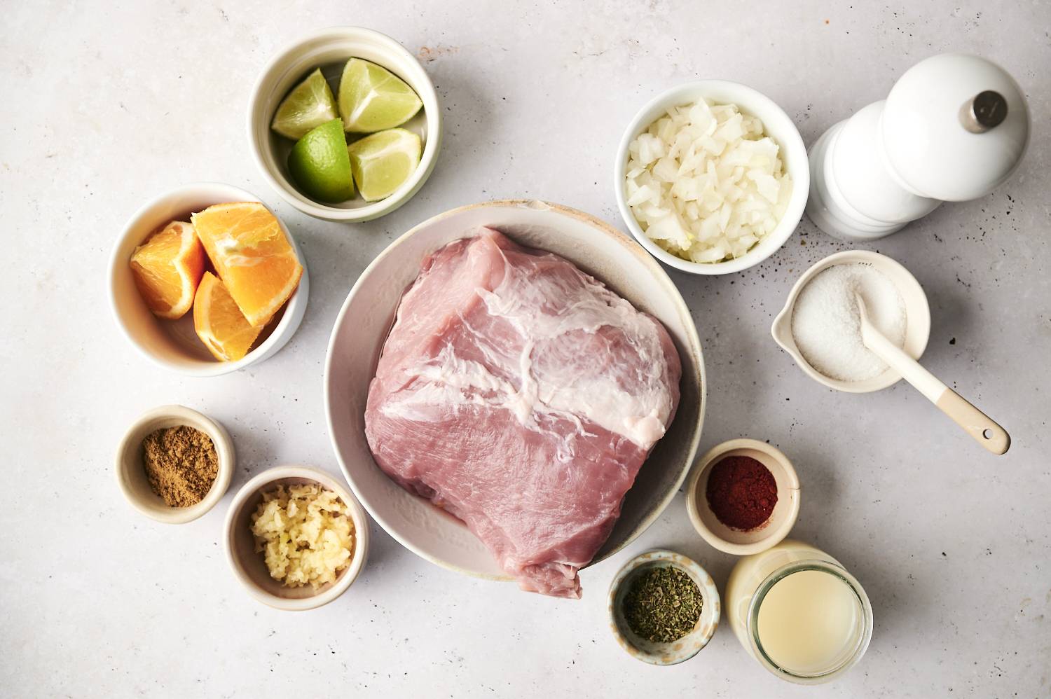 Raw pork shoulder in a bowl surrounded by ingredients including lime wedges, orange slices, chopped onions, minced garlic, spices, herbs, and chicken broth