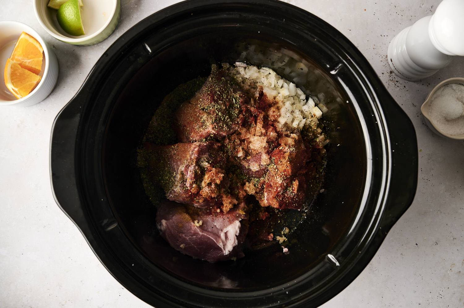 A slow cooker filled with raw pork, chopped onions, and various spices, surrounded by small bowls containing orange and lime wedges, salt, and seasoning.