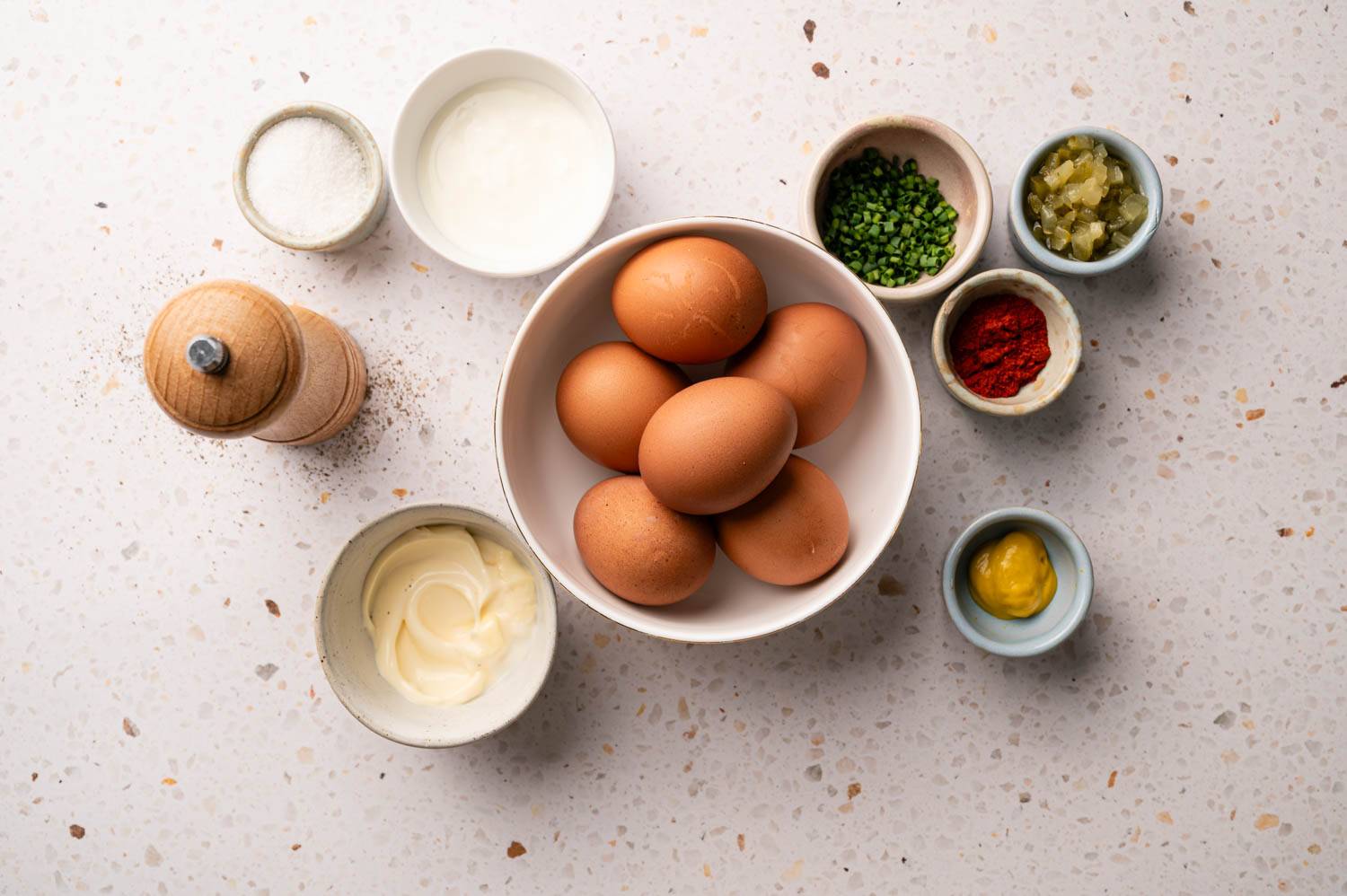 A white countertop features a bowl of brown eggs surrounded by bowls of salt, yogurt, chives, relish, paprika, mustard, and mayonnaise, with a pepper grinder.