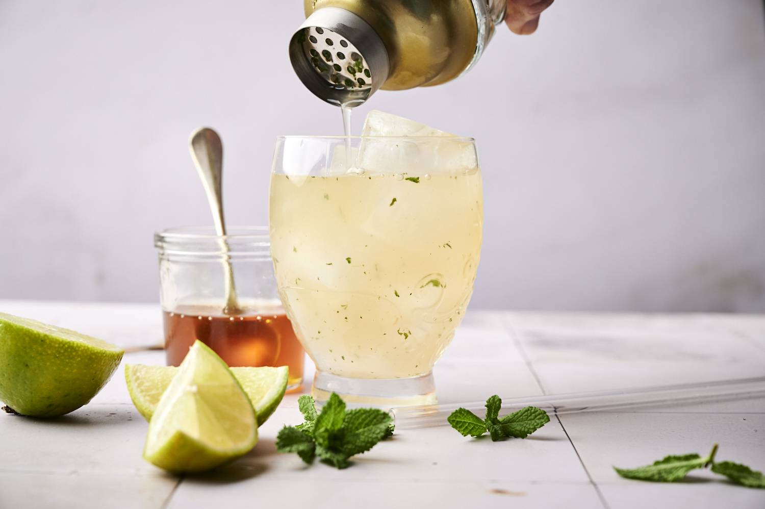 A refreshing cocktail being poured over ice in a glass, surrounded by lime wedges, mint leaves, and a jar of honey.
