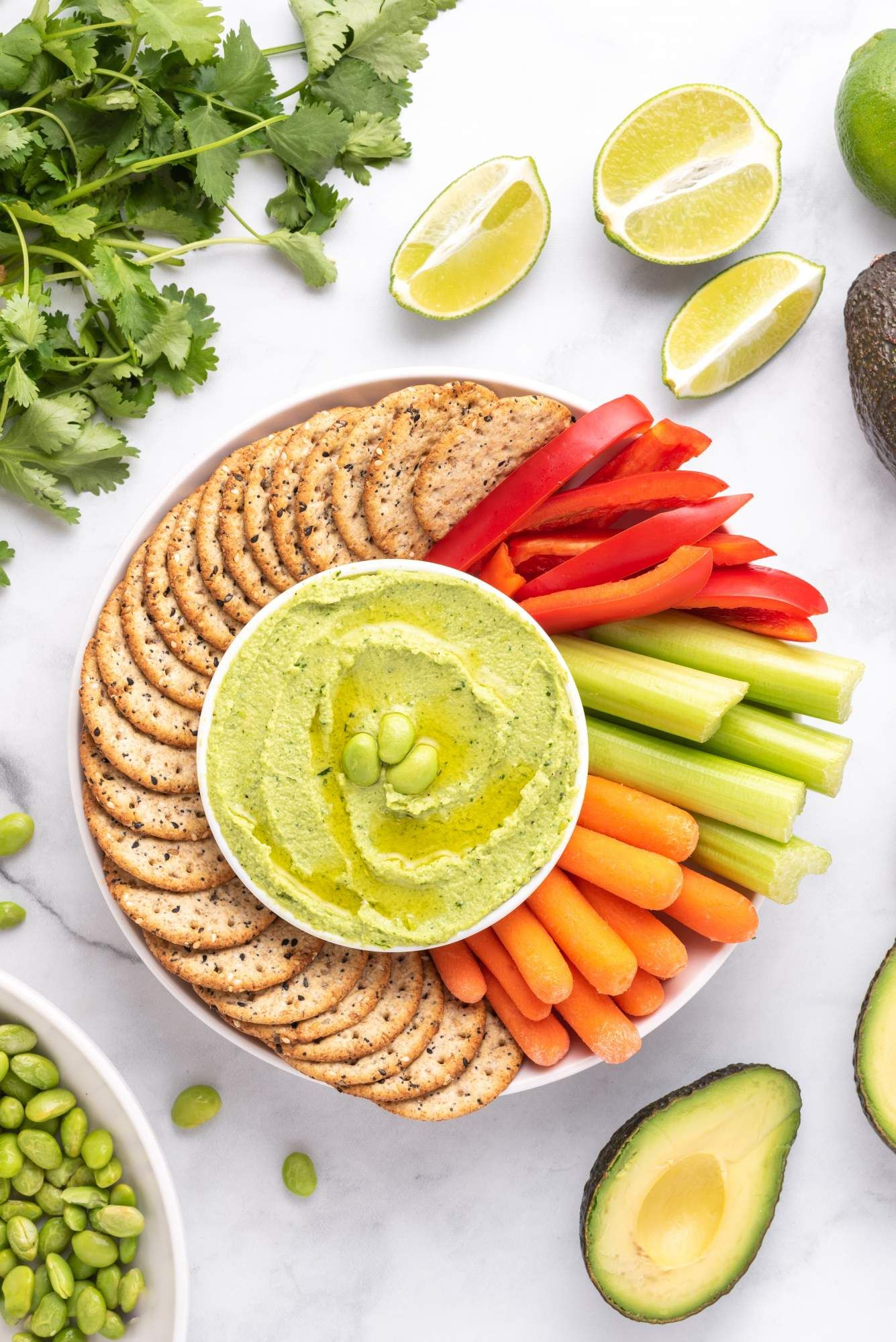 Cilantro lime edamame hummus in a bowl with carrots, red peppers, celery, and crackers on the side.