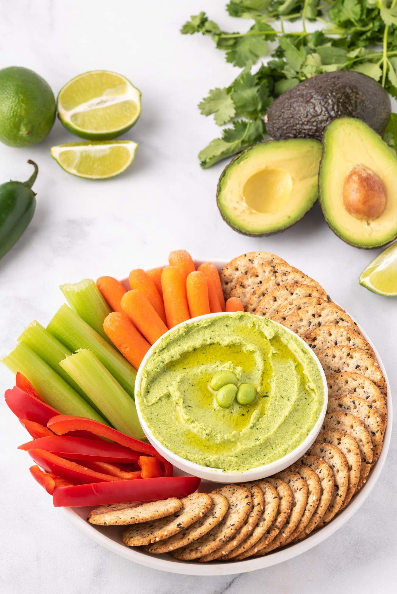 Hummus made with edamame with avocado, cilantro, edamame, and fresh vegetables on the side for dipping.