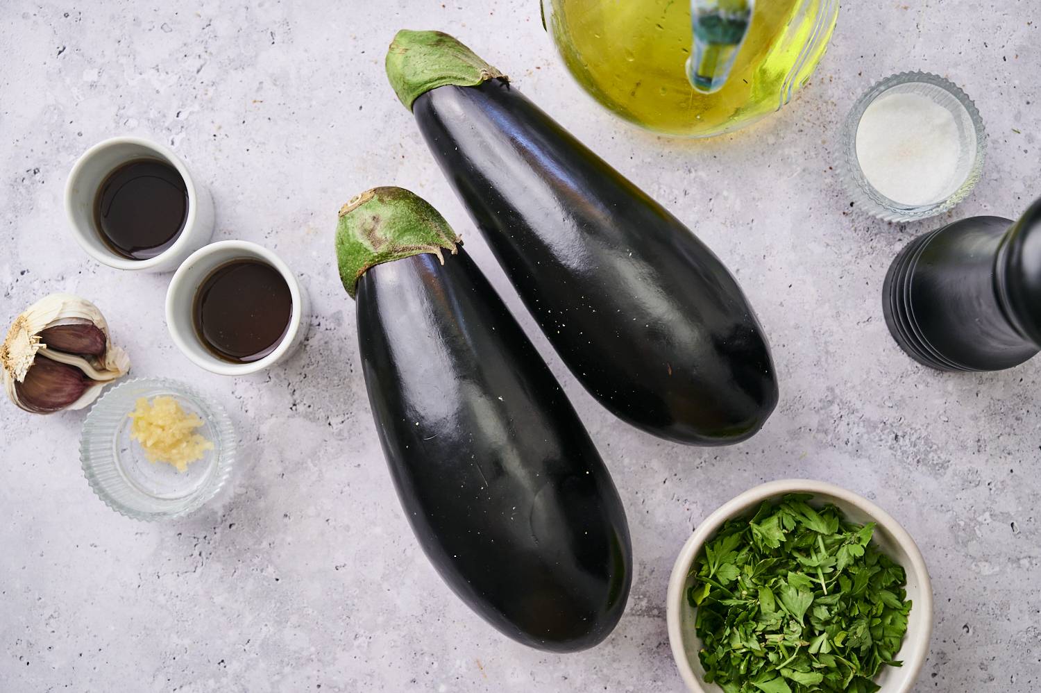 Two glossy eggplants, garlic, ginger, two types of sauces, olive oil, salt, pepper, and fresh herbs on a grey surface.