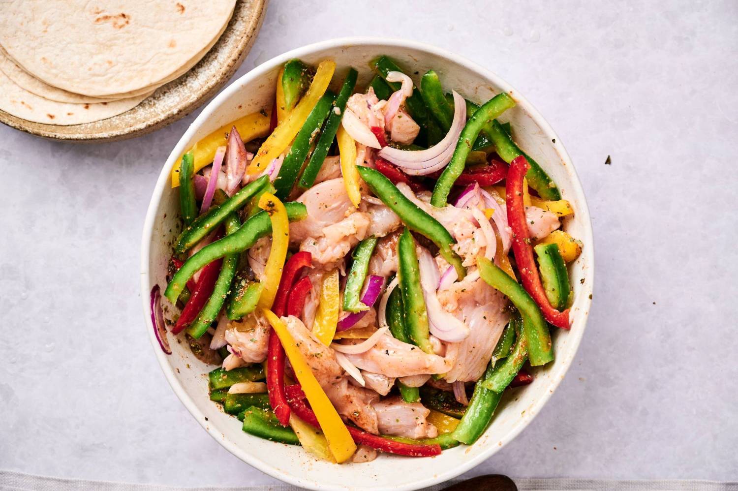 Chicken breast, bell peppers, and red onions tossed with olive oil, fajita seasoning, and lime juice in a bowl.