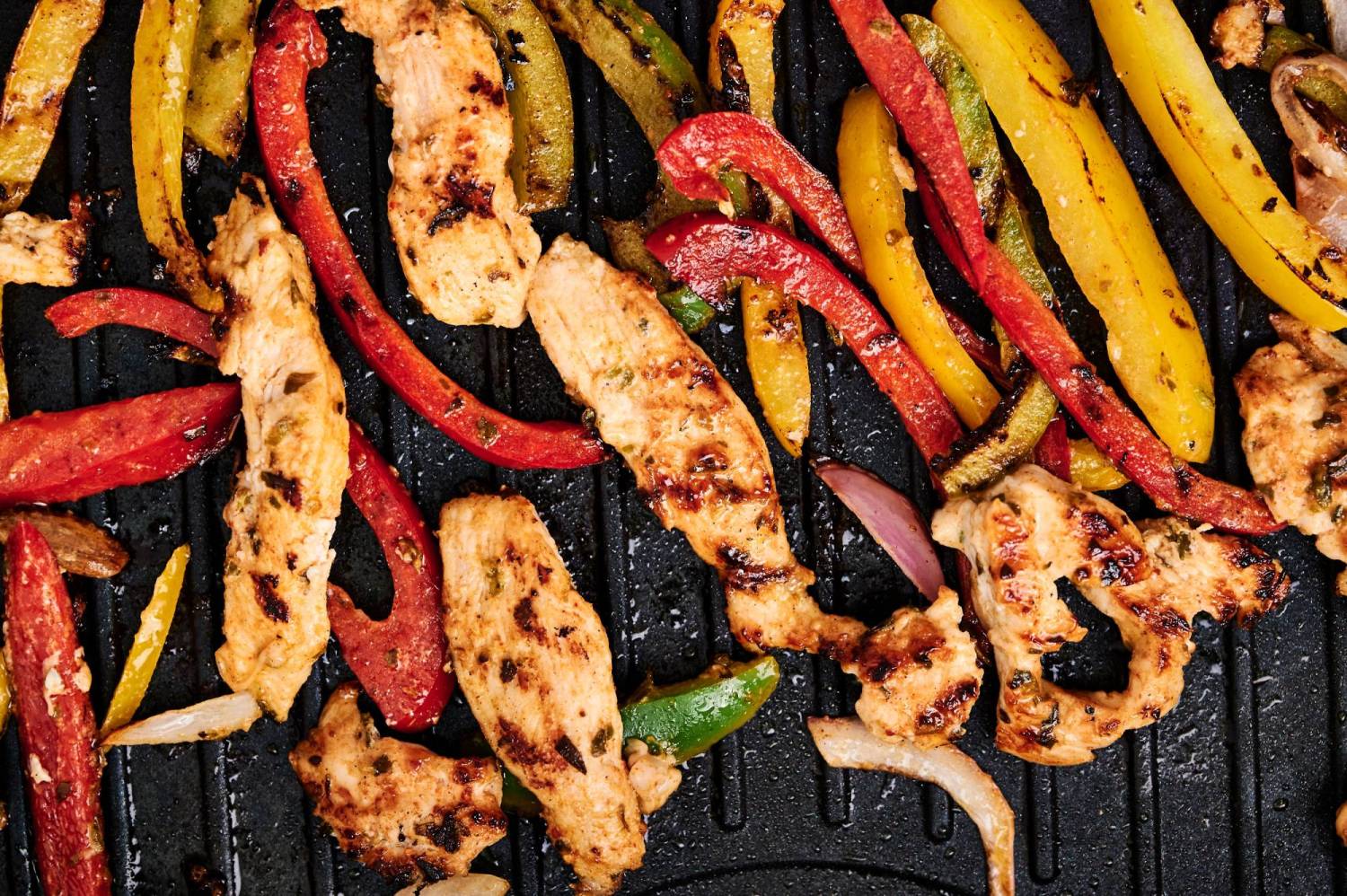 Chicken fajitas with bell peppers and onions cooking on a grill pan.