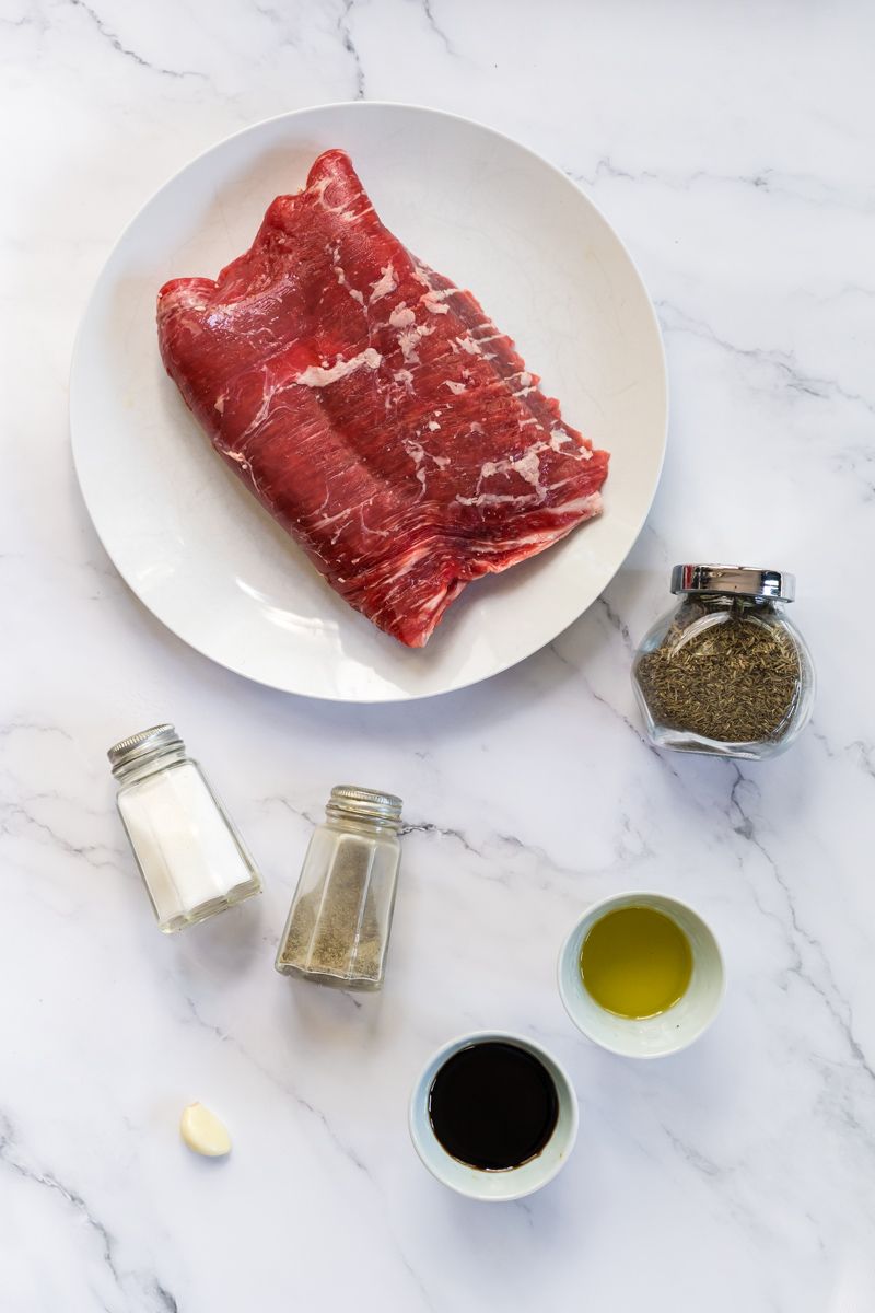 Ingredients for balsamic grilled flank steak including raw steak, garlic, balsamic vinegar, thyme, salt, and pepper.