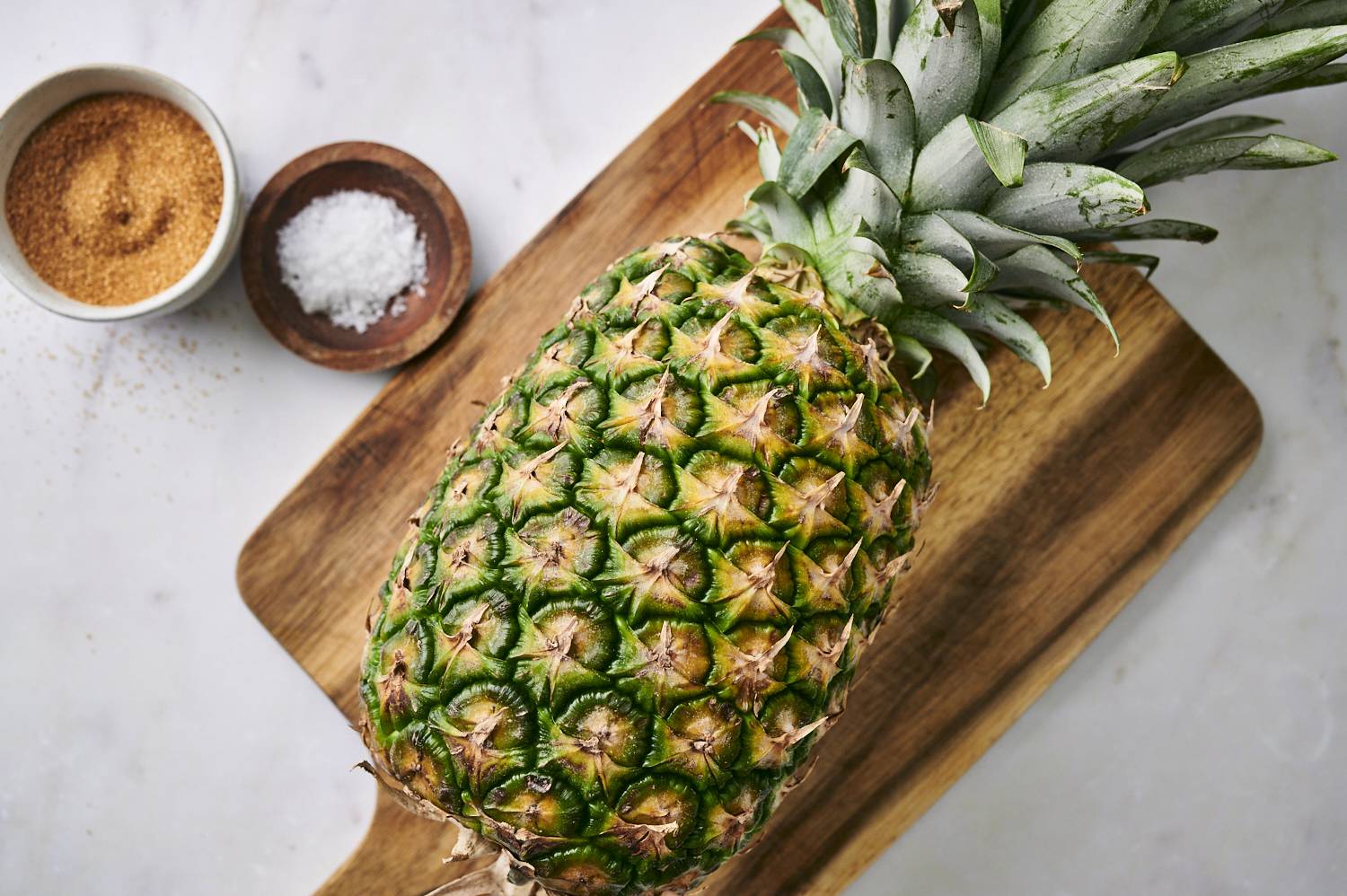 A fresh pineapple on a wooden cutting board, accompanied by bowls of brown sugar and sea salt.