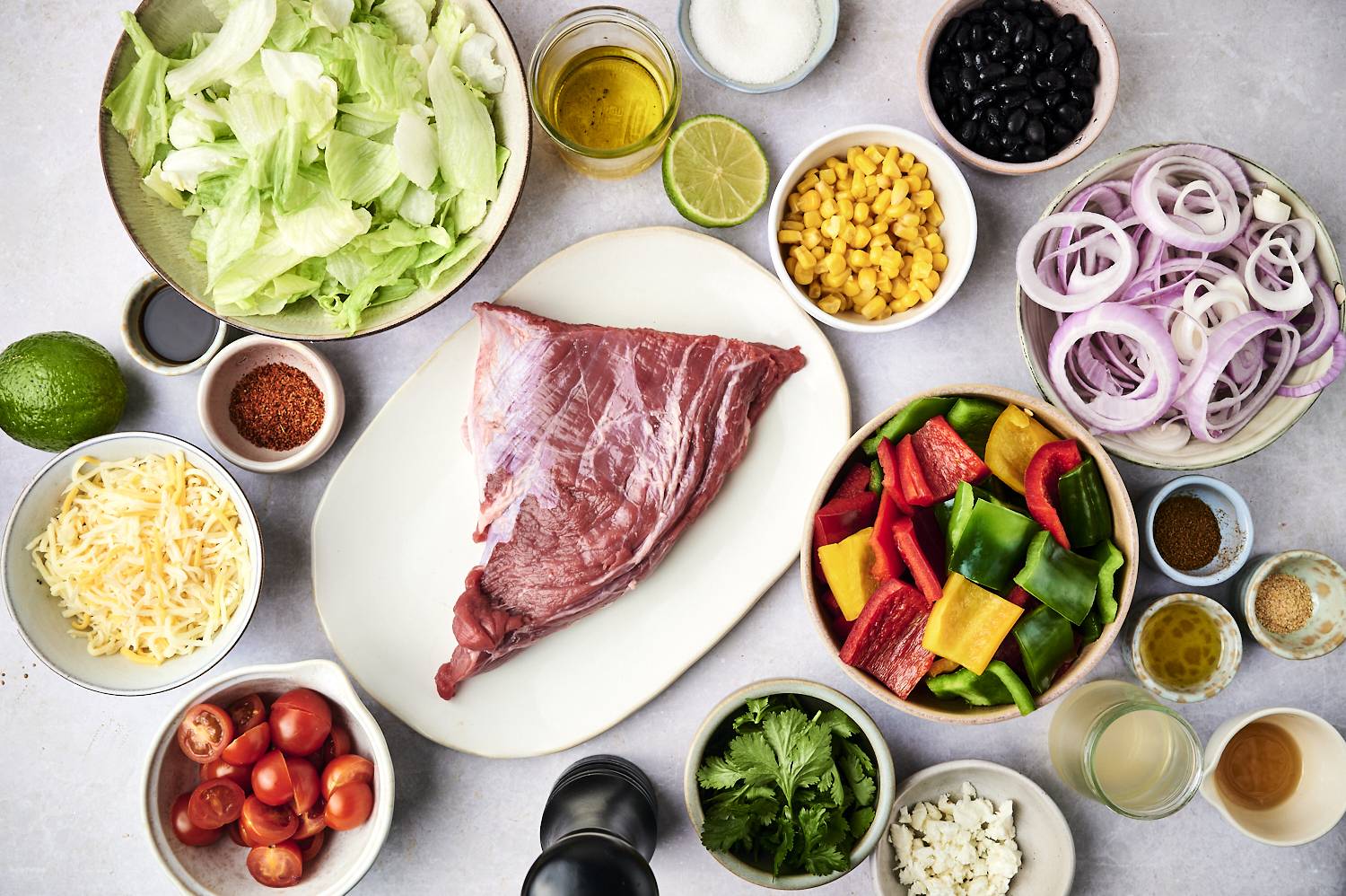 An assortment of fresh ingredients for a salad: lettuce, tomatoes, bell peppers, black beans, corn, cheese, onions, and uncooked beef.