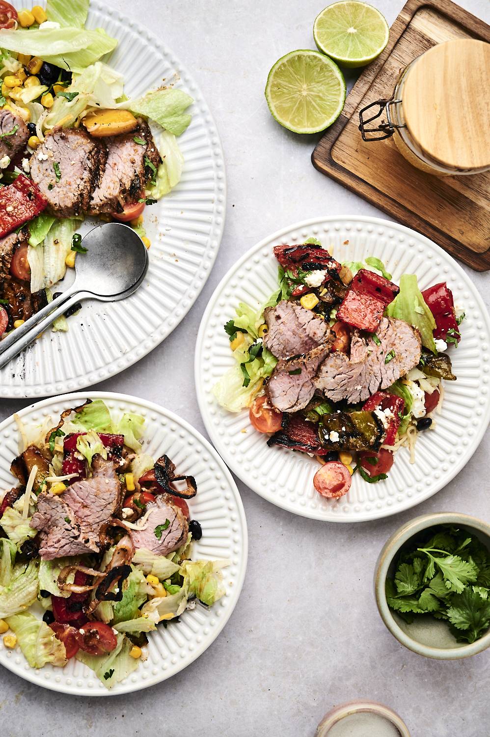 Three vibrant plates of salad topped with sliced beef, lettuce, tomatoes, and corn, with lime and a jar of dressing in the background.