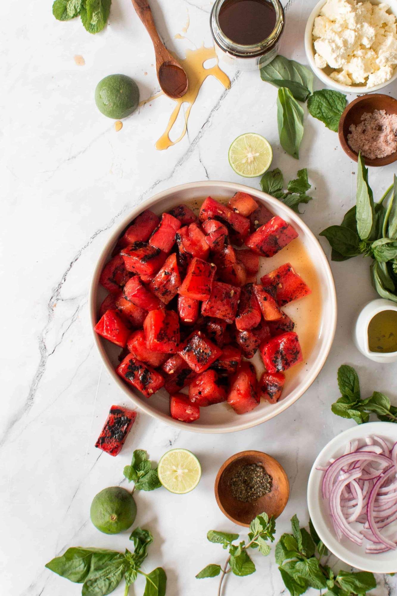 Watermelon, feta, and mint salad with grilled watermelon, limes, and herbs on a marble board.