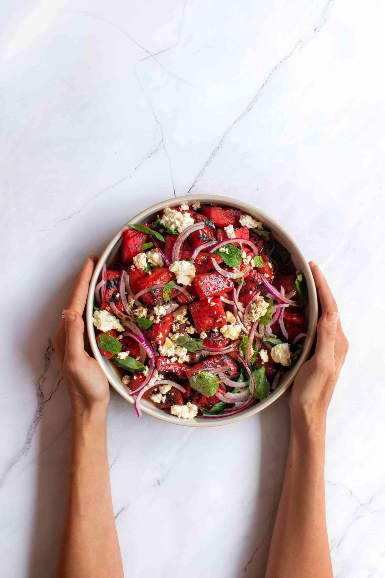Grilled watermelon, feta, and herb salad with red onions in a bowl with two hands holding it. 