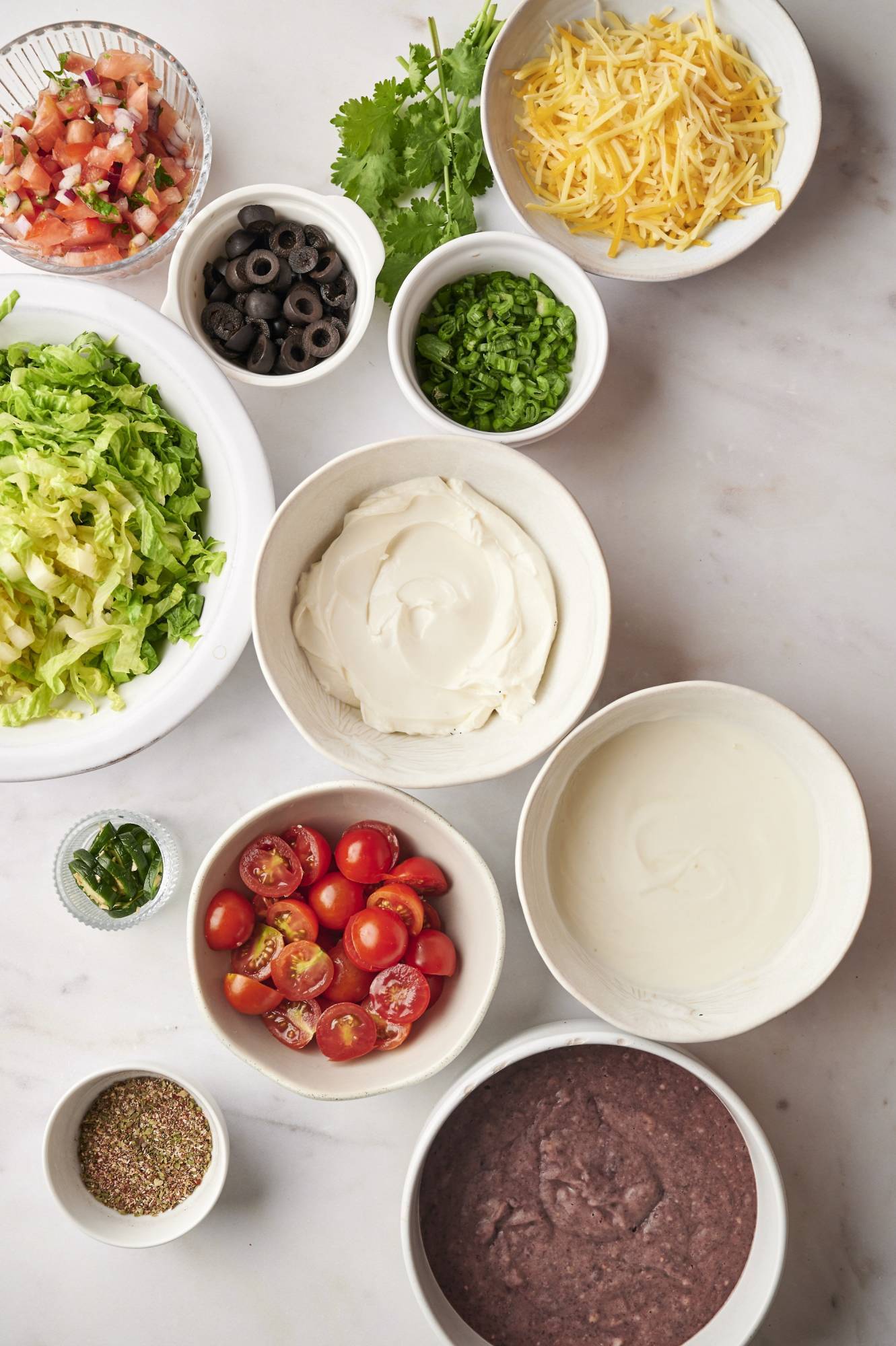 Ingredients for healthy taco dip including light cream cheese, Greek yogurt, refried beans, salsa, lettuce, shredded Mexican cheese, olives, jalapenos, and lettuce.