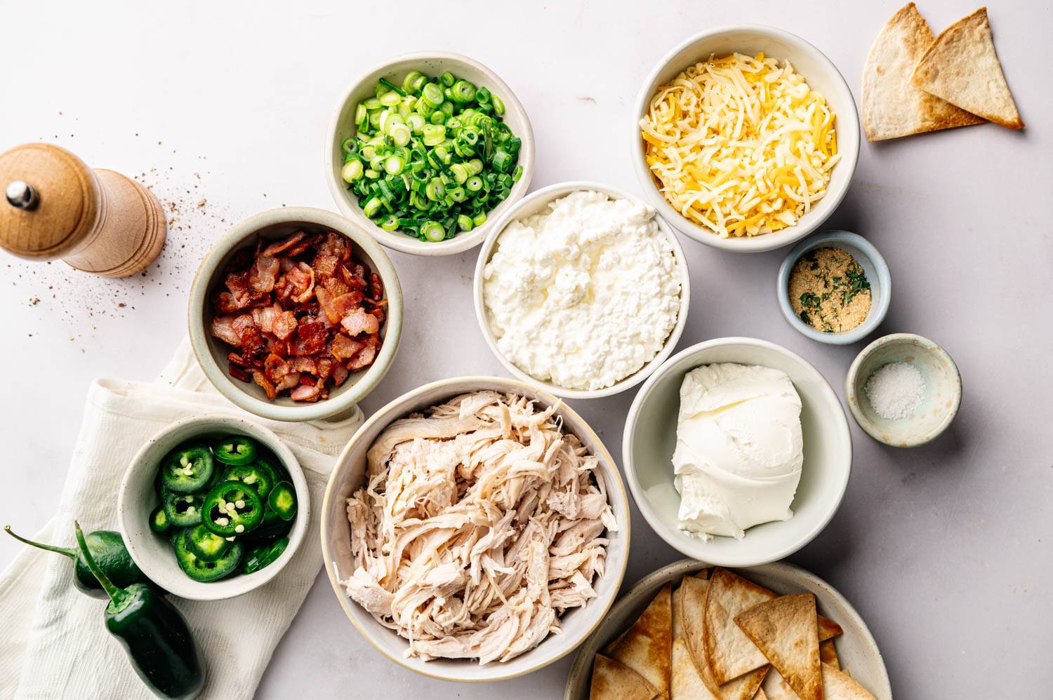 Bowls of ingredients for a flavorful dish include shredded chicken, bacon, green onions, jalapeños, shredded cheese, cottage cheese, cream cheese, herbs, and salt, with tortilla chips on the side.