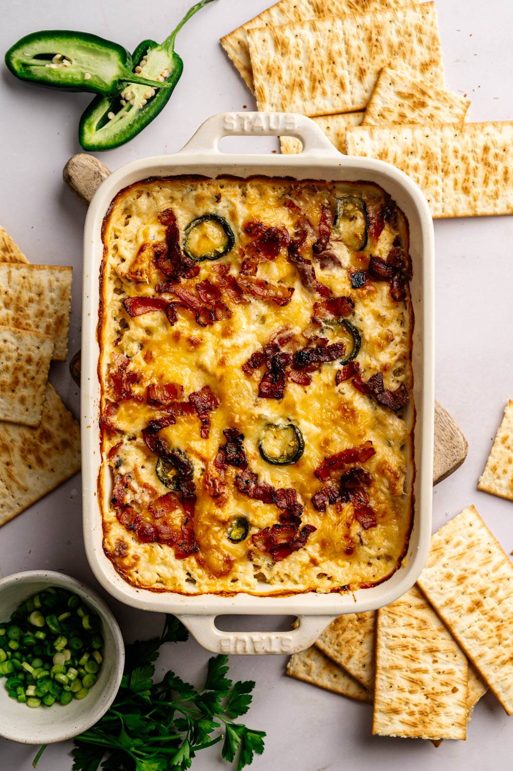 Baked casserole topped with melted cheese, bacon, and jalapeño slices, surrounded by flatbread crackers. A small bowl of chopped green onions and parsley garnish beside.