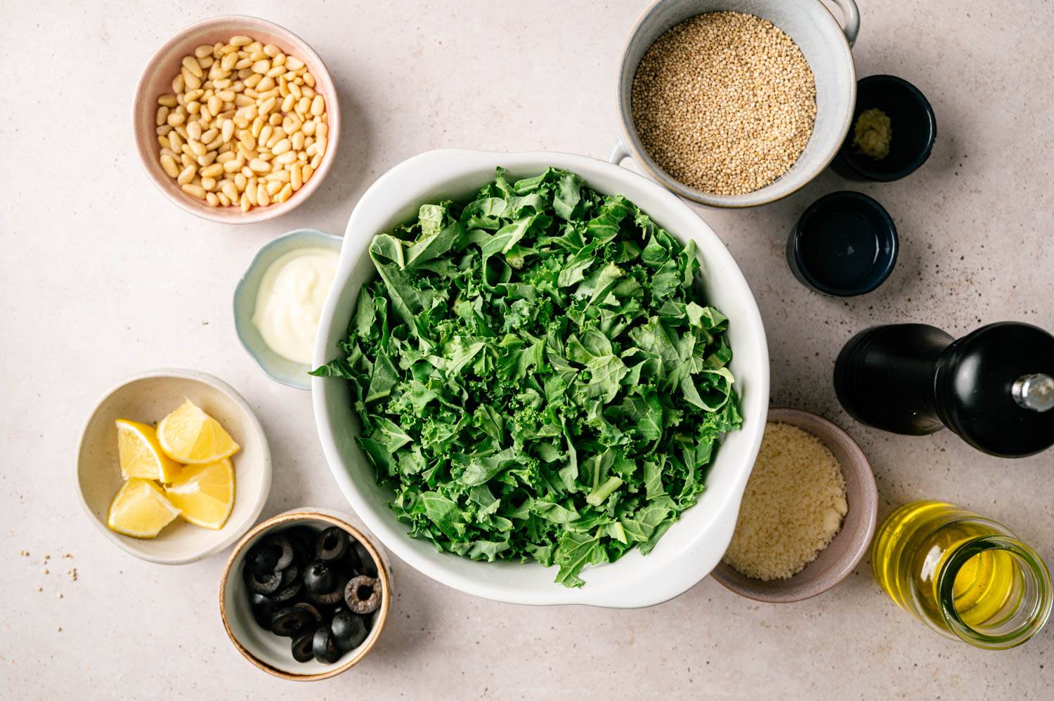 A white bowl of fresh kale is surrounded by ingredients: pine nuts, lemon wedges, black olives, quinoa, parmesan, oil, and creamy dressing, evoking a fresh, healthy vibe.