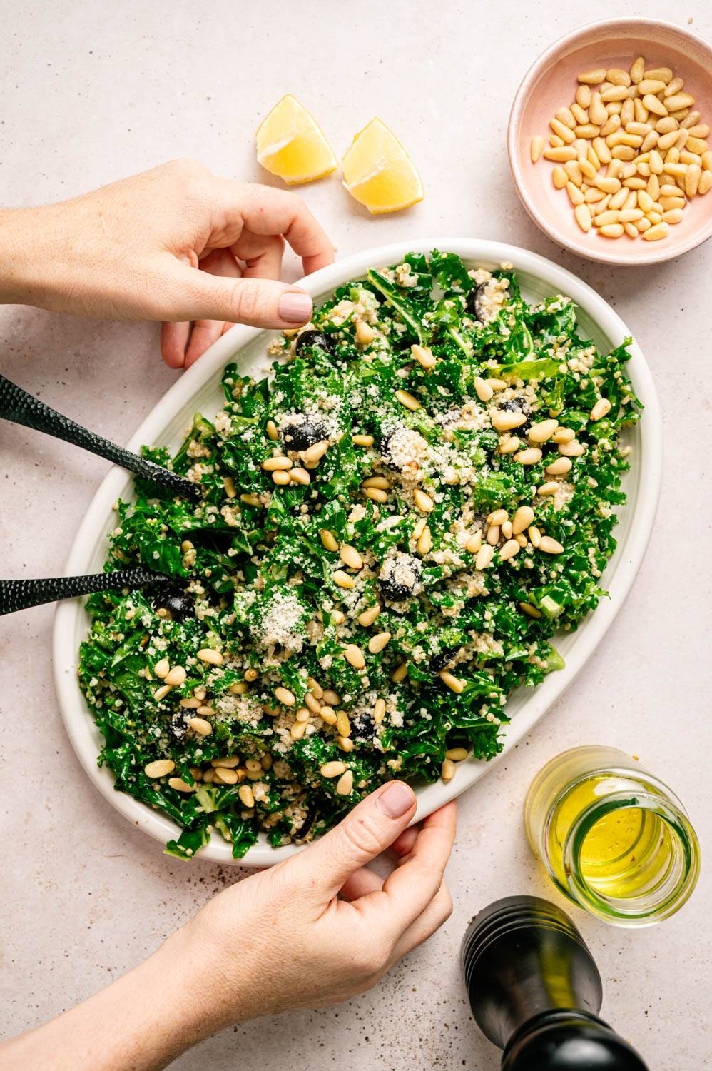 A fresh kale salad with pine nuts and grated cheese on an oval plate. Two lemon wedges, a bowl of pine nuts, and olive oil are nearby. Hands adjust the plate.