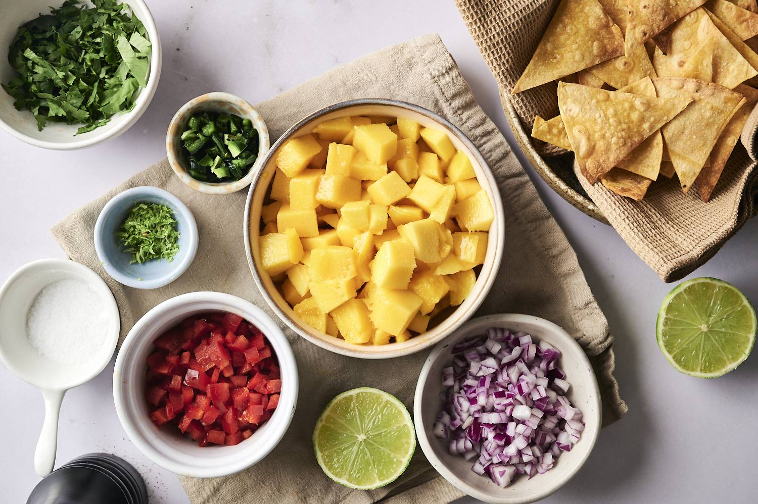 Overhead view of fresh mango salsa ingredients in bowls, including diced mango, red onion, red bell pepper, jalapeño, lime zest, cilantro, and a basket of crispy tortilla chips.